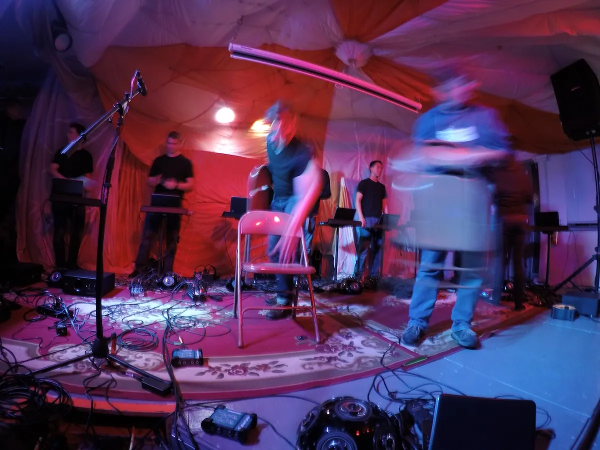 Wide-angle view of an experimental electronic music performance — figures at keyboards and laptops under red-draped fabric, with motion blur and coloured stage light.