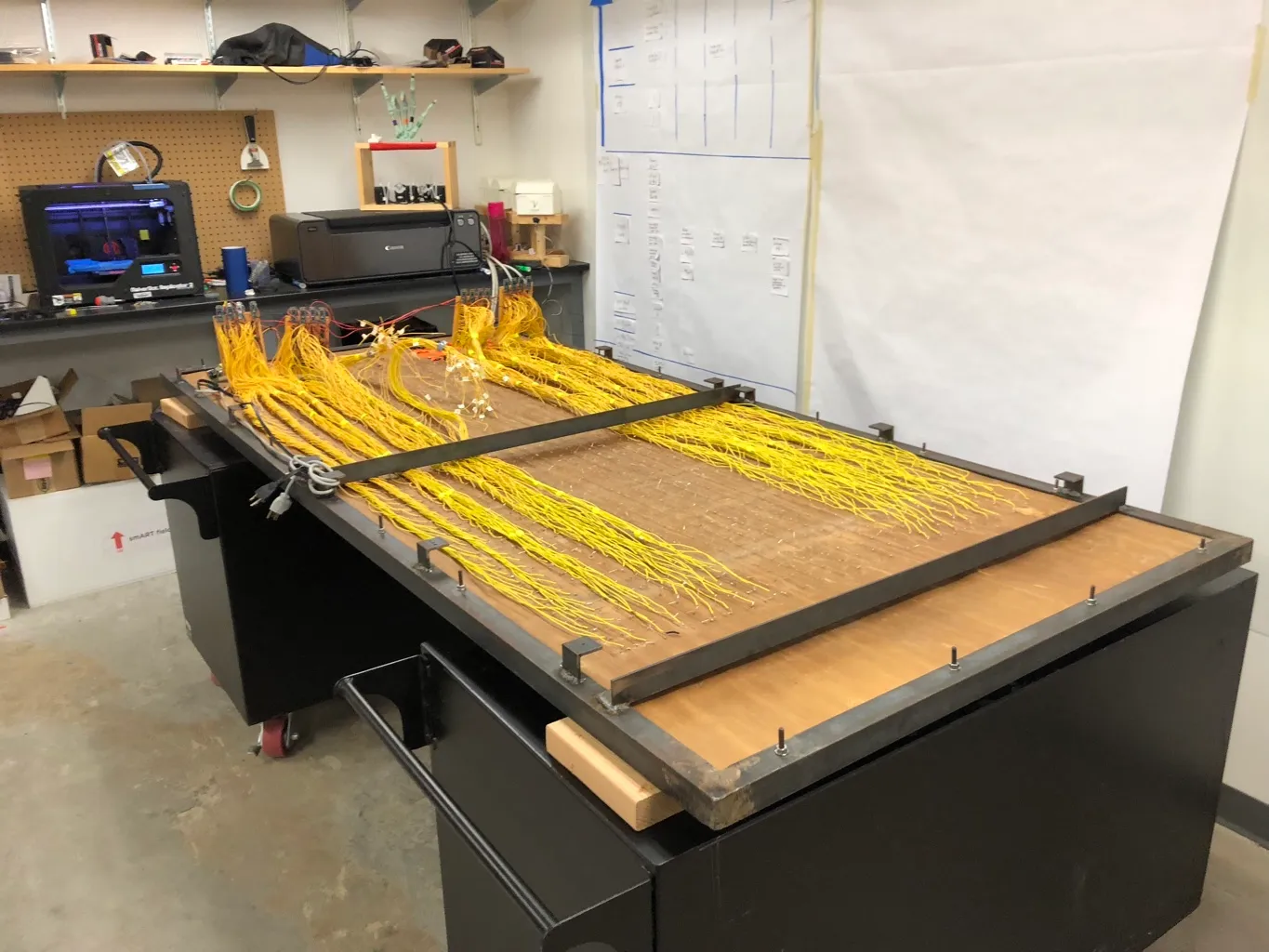 A large wooden panel embedded with rows of small LEDs and hundreds of yellow wires lies on a black metal fabrication table in a workshop, with the wires gathered into bundles feeding toward control electronics. A 3D printer, printer, and whiteboards with diagrams are visible in the background.