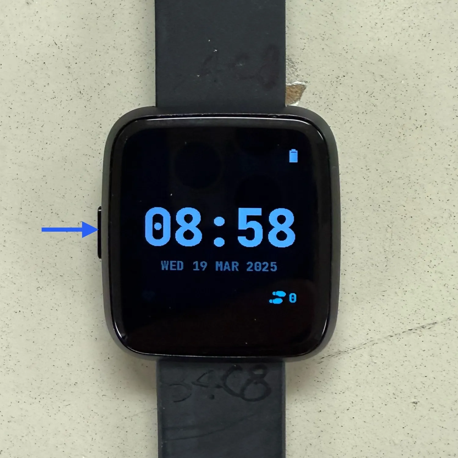 A black square-faced smartwatch on a black silicone band, photographed against a textured beige wall. The screen displays the time 08:58 in blue digits, the date "WED 19 MAR 2025," a battery icon, and a step counter reading 0. A blue arrow points to a physical button on the left side of the watch case.